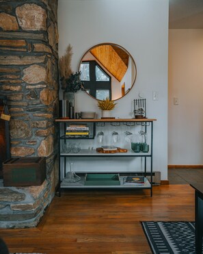 A room with a stone wall, a wooden floor, a mirror, a shelf with books and glasses, and a small table with a vase and a book.