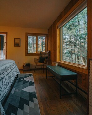 A room with a bed, a chair, a table, and a large window offering a forest view.