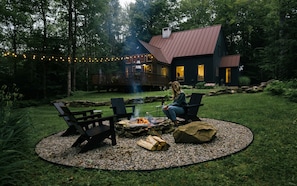 Un jardin avec un foyer, deux fauteuils Adirondack et une terrasse menant à une maison dont le toit est en métal.