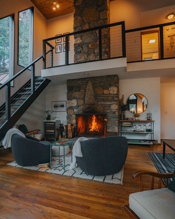 A spacious living room with a stone fireplace, comfortable seating, a staircase, and a view of the outdoors.