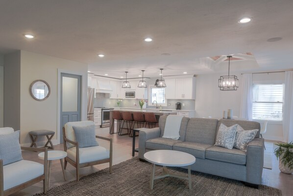 A modern living room with a grey sofa, two armchairs, and a coffee table. A kitchen with white cabinets and pendant lighting is visible in the background. - Lakehouse Getaway - Grand Rapids Area - Murray Lake - Sandy Beach, Dock, Hot Tub (Lowell)
