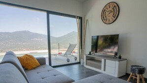 A modern living room with a grey sofa, yellow cushions, a flat-screen TV, a clock, and a view of a beach and mountains through large windows. - Villa Cupabia, sea view and heated pool (VIGGIANELLO)
