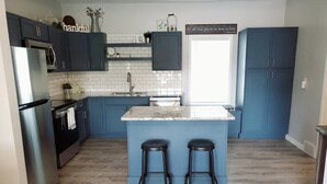 A modern kitchen with dark blue cabinets, a white island with two stools, a stainless steel refrigerator, and a window with a view. - Dog Friendly|Fenced|Full Kitchen|Firepit (Alpena)