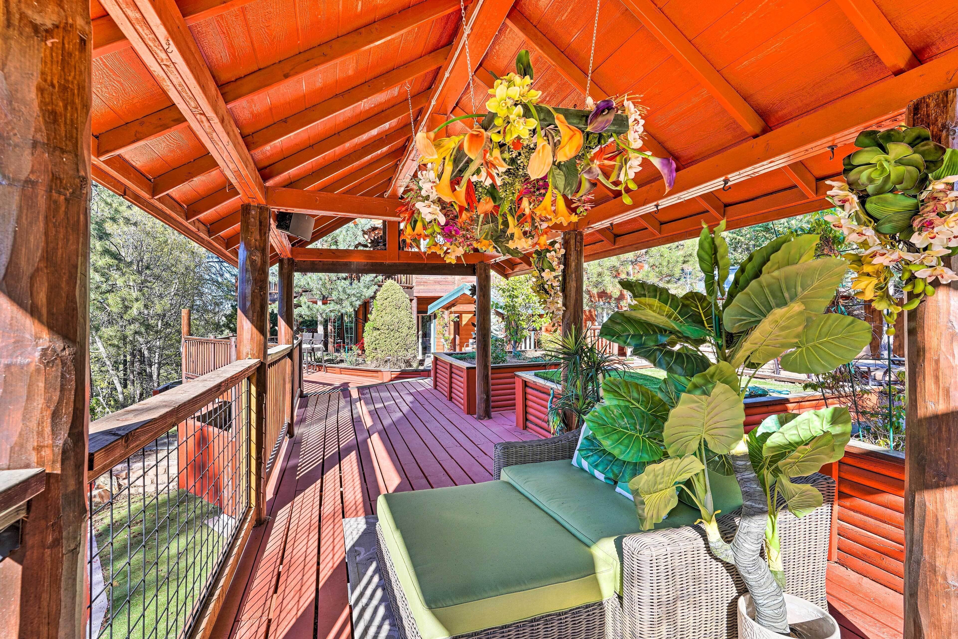 Una terraza de madera con un arreglo floral colgante, un sofá de mimbre y vistas de la vegetación circundante.