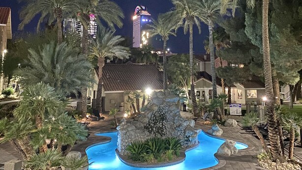 Vista noturna de um resort com piscina cercada por palmeiras e um prédio com sinalização iluminada.