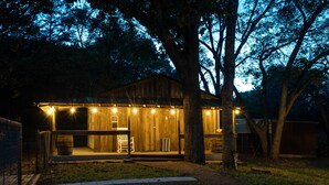 A wooden cabin with a porch, illuminated by string lights, surrounded by trees. - Secluded Country Retreat- Hot Tub & Fire Pit-3BR minutes to Z-Plex and Downtown  (McKinney)