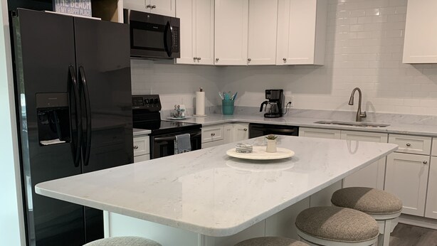 Una cocina moderna con gabinetes blancos, un refrigerador negro, horno empotrado, microondas, cafetera y un lavamanos. Hay cuatro taburetes con asientos acolchados en la isla.