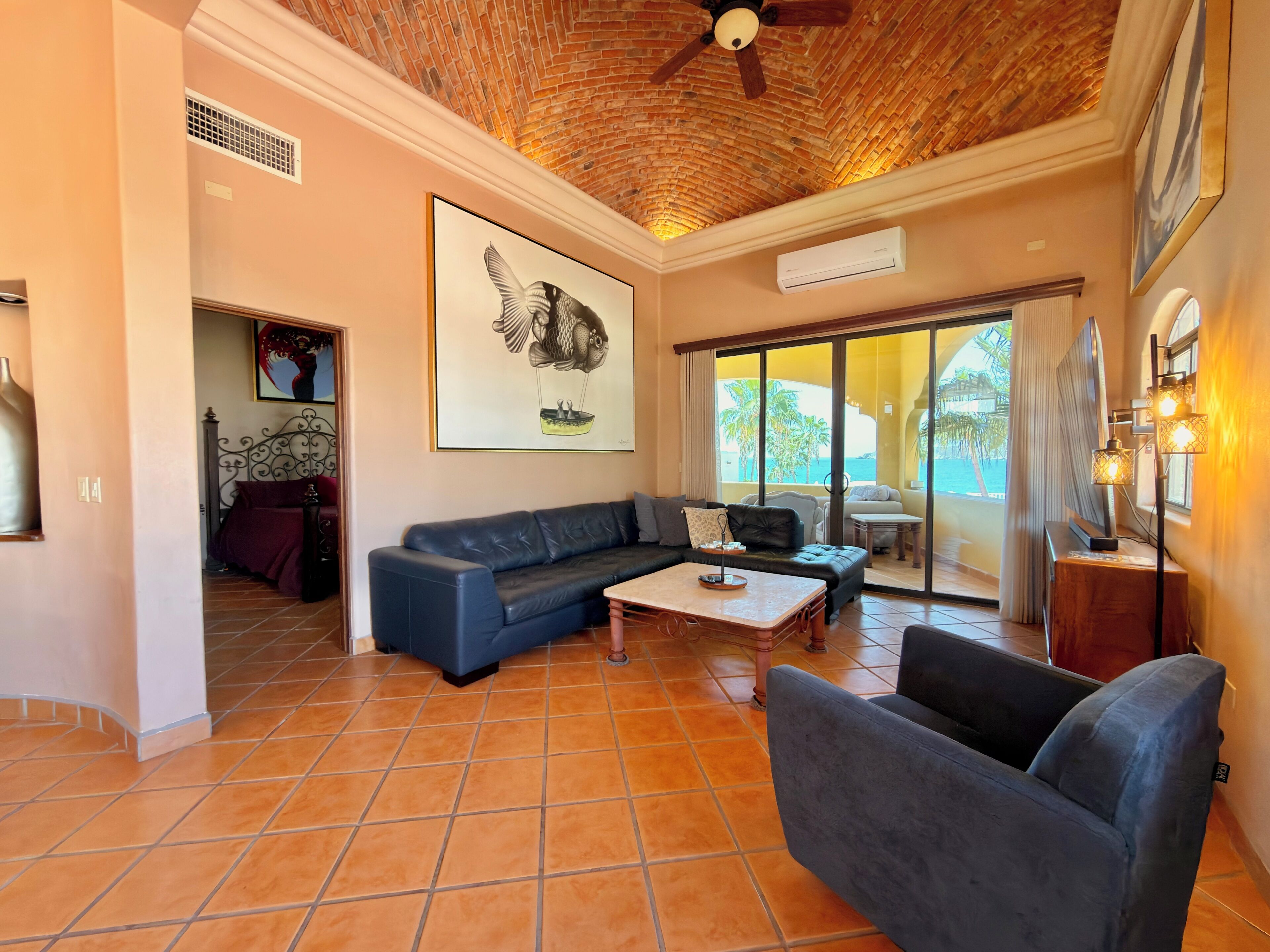 A living room with a sectional sofa, a coffee table, and a ceiling fan. There is a large window with a view of palm trees and a pool.
