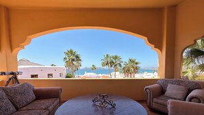 Una habitación con una mesa redonda de madera, dos sofás con diseños, y un gran ventanal con arco que da a una playa con palmeras y un cielo azul despejado.