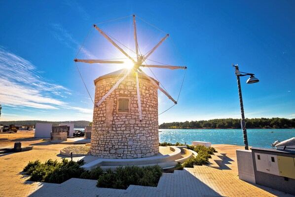 A stone windmill with a circular base and a metal roof, surrounded by a paved area with benches and a lamp post, overlooking a body of water.