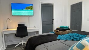 Un dormitorio con cama, escritorio con computadora, televisor en la pared y dos puertas.