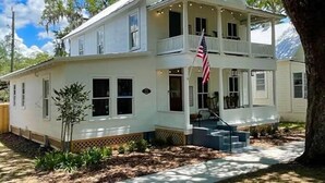 Una casa blanca de dos pisos con un porche, una bandera y un árbol en primer plano.