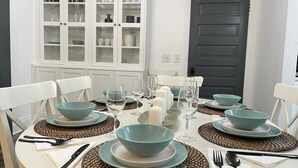 Una mesa de comedor dispuesta con fuentes, platos y cubiertos en color aguamarina, rodeada de sillas blancas y un armario blanco con puertas de cristal.