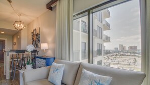 Een moderne woonkamer met een beige bank, kussens met stervormige patronen, een kroonluchter, een eethoek met een tafel en stoelen, en een groot raam met uitzicht op de stad.