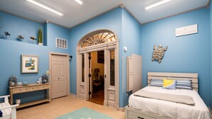 A bedroom with a bed, a desk, a chair, a dresser, and a door leading to another room.