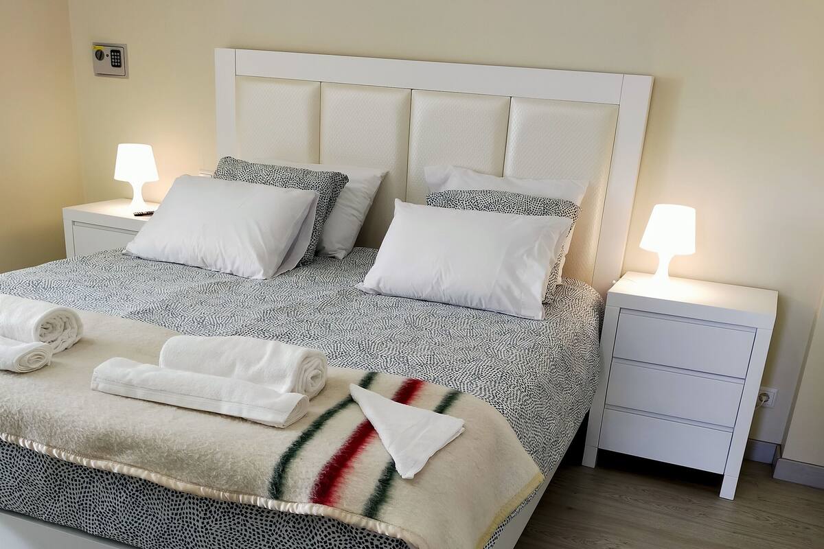 Una cama bien hecha con una colcha estampada, dos almohadas blancas y un edredón con un listón rojo. Hay dos mesitas de noche con lámparas, y una mesita de noche blanca con un cajón.