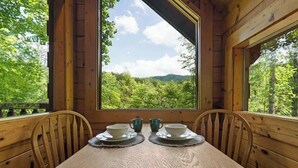 Ein Holztisch mit zwei Stühlen, ein Fenster mit Blick auf einen Wald und zwei Tassen auf dem Tisch.