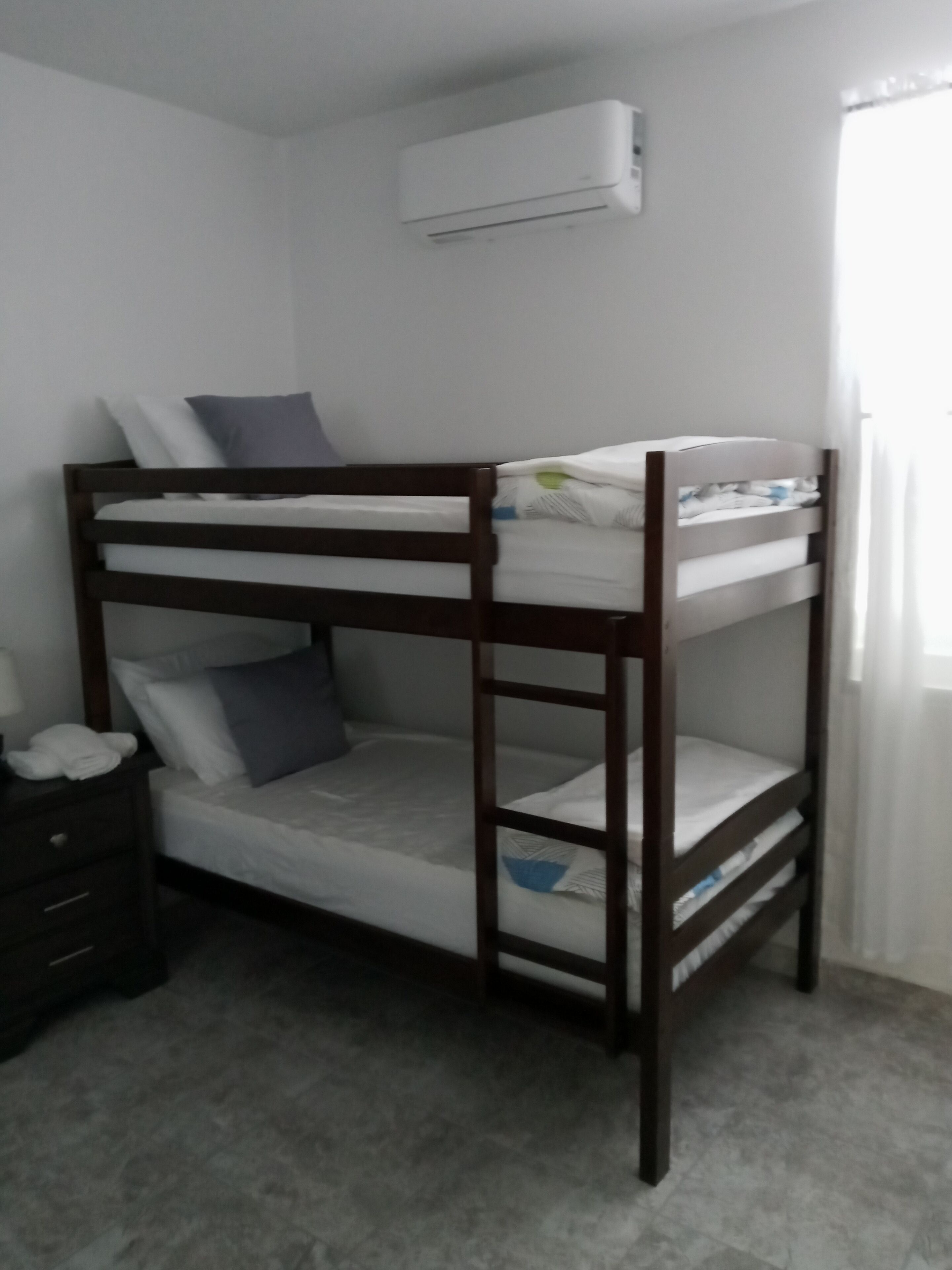 Un lit superposé dans une chambre où l’on aperçoit un climatiseur blanc fixé au mur et une table de chevet avec une lampe.
