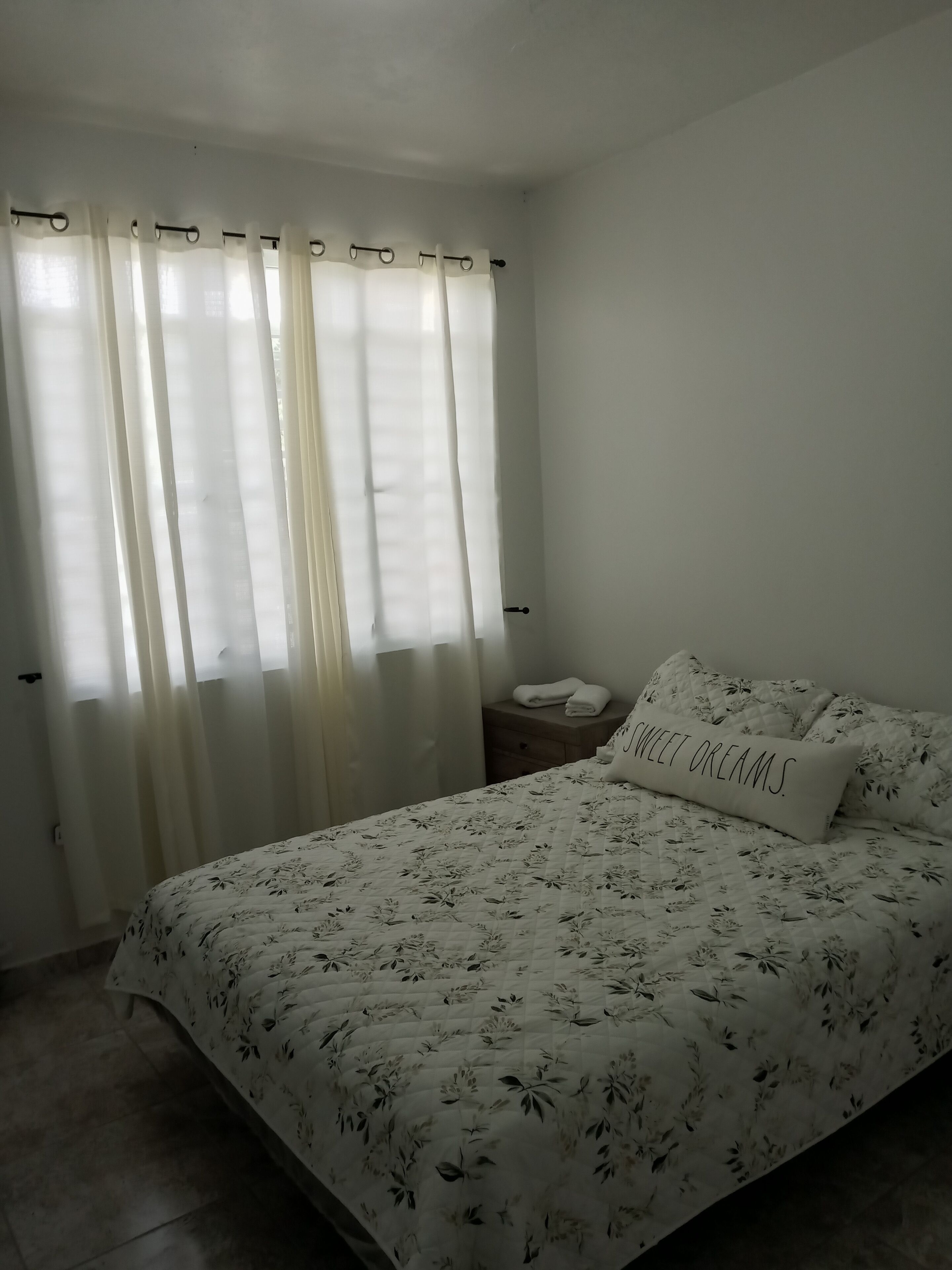 Une chambre à coucher avec un lit, une table de chevet, un oreiller orné d’un motif, et une fenêtre avec des rideaux.