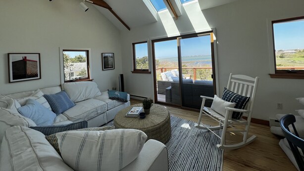 A spacious living room with a high ceiling, skylights, a large sliding glass door leading to an outdoor area, a white sofa, a rocking chair, and a coffee table. - Chappaquiddick Getaway with stunning Ocean Views, walk to the beach (Edgartown)