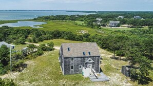 A large, rectangular stone building with a gabled roof, surrounded by lush greenery and a body of water in the background. - Chappaquiddick Getaway with stunning Ocean Views, walk to the beach (Edgartown)