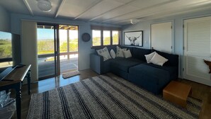 A spacious living area with a dark blue sofa, a wooden coffee table, and a large rug. There is a sliding glass door leading to an outdoor deck. - Chappaquiddick Getaway with stunning Ocean Views, walk to the beach (Edgartown)