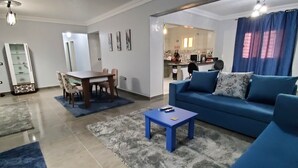Amplia sala de estar con un sofá azul, una mesa de comedor con sillas y una cocina al fondo.