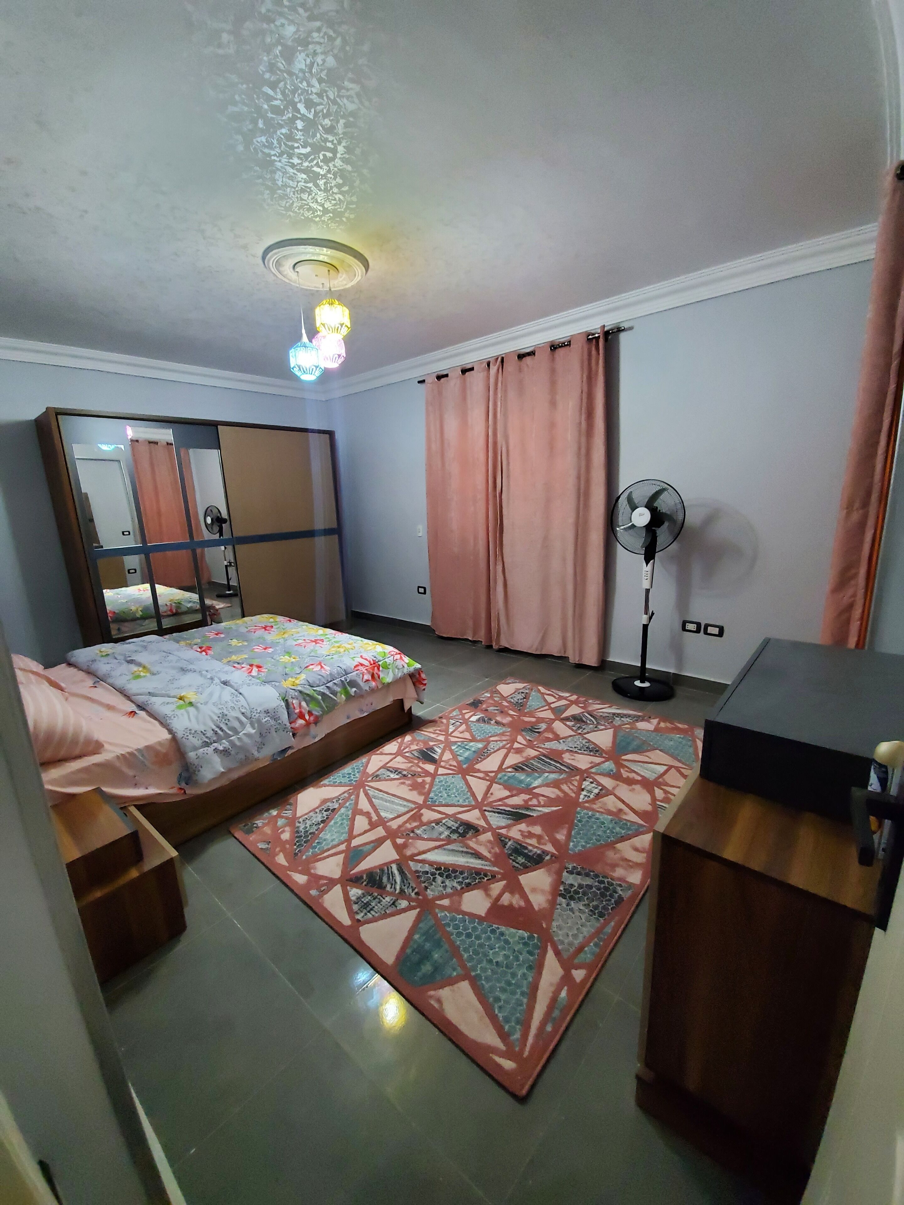 Un dormitorio con cama, escritorio, ventilador de pie, alfombra estampada y lámpara del techo.