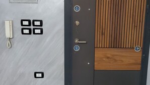 En moderne dør med en kombination af træ- og metalpaneler, et nøglehul og en lås. Der er et intercom og et telefonapparat monteret på væggen til venstre.