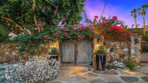 Una entrada de paredes de piedra con un portón de madera, rodeada de abundante follaje y plantas florecientes.