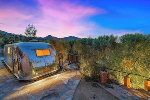 A vintage camper parked on a stone pathway with a wooden deck and metal railing, surrounded by dense shrubbery and mountains in the background. - Jaguar Villa - 6 BR in the Palm Desert hills! (Palm Desert)