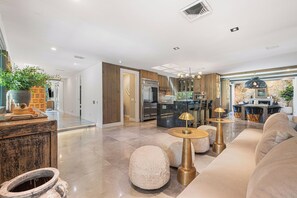 Eine moderne Küche mit Backofen, ein Essbereich mit einer Chandelüster und ein Wohnzimmer mit einer Couch und runden Hocker.