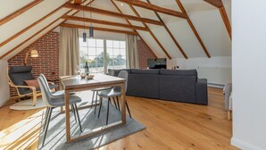 Un loft espacioso con techo alto, vigas de madera a la vista, una zona de comedor con mesa y sillas, un sofá gris y una pared de ladrillo.