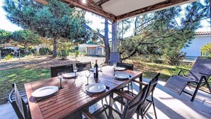 An outdoor dining area with a wooden table set for two, surrounded by chairs, under a covered roof with a view of a garden and trees. - Stunning private villa for 6 guests with WIFI, TV, terrace and pets allowed (Montalivet-les-bains)