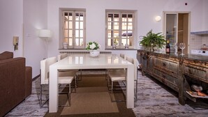 Een moderne eethoek met een witte tafel en stoelen, twee ramen met luiken, een houten kast met spiegel en een kleine plant op de vensterbank.