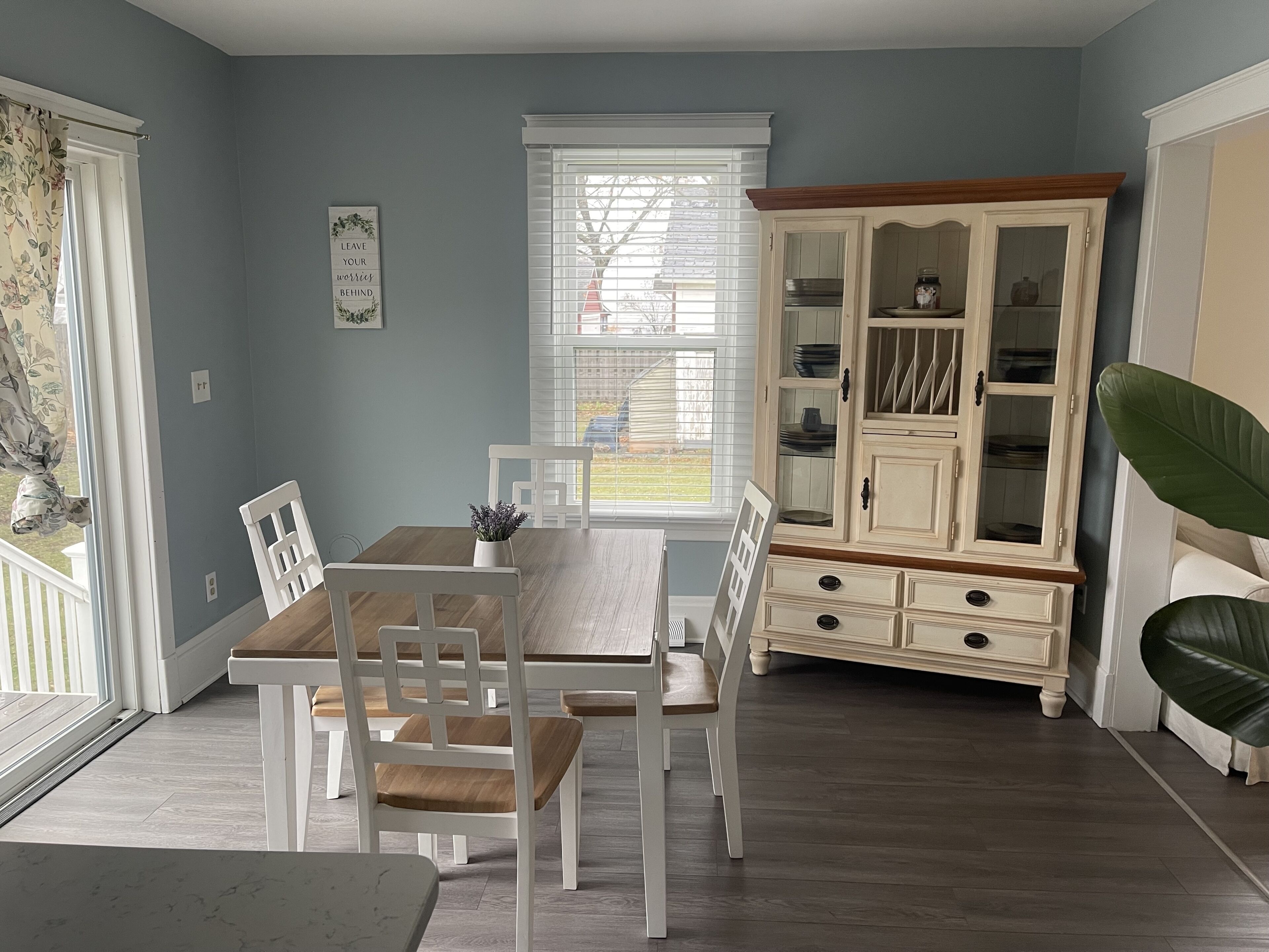 A dining area with a wooden table and white chairs, a white cabinet with glass doors, and a large window with blinds.