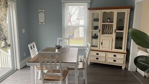 Un coin repas avec une table en bois et des chaises blanches, un buffet blanc aux portes vitrées et une grande fenêtre avec des stores.