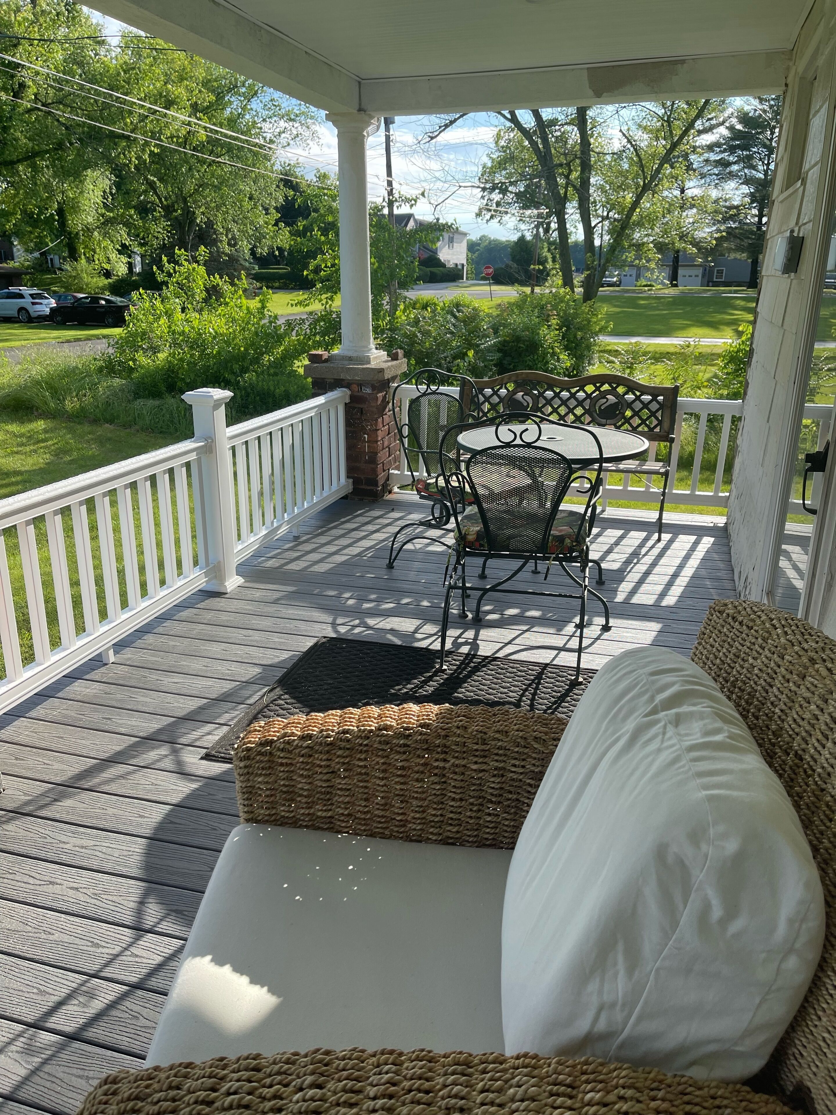 A covered porch with a white railing, a wicker sofa with a white cushion, and a metal table with chairs.