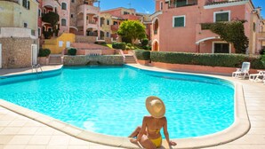 Une zone autour d’une piscine où l’on voit une personne assise au bord, entourée de bâtiments dotés de balcons et d’escaliers menant à la piscine.