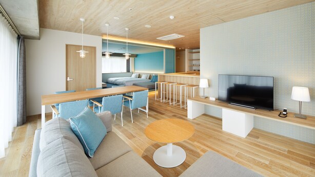 A modern living room with a sofa, dining table, chairs, a TV, and a kitchen area in the background.