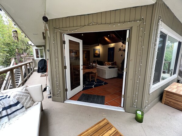 A covered outdoor area with a wooden deck, a seating area with a bench, and a view of the interior through an open door.