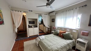 A bedroom with a bed, nightstands, a desk, a chair, a ceiling fan, and a window with curtains. - Hideaway Steps to Beaches of Leucadia (Encinitas)