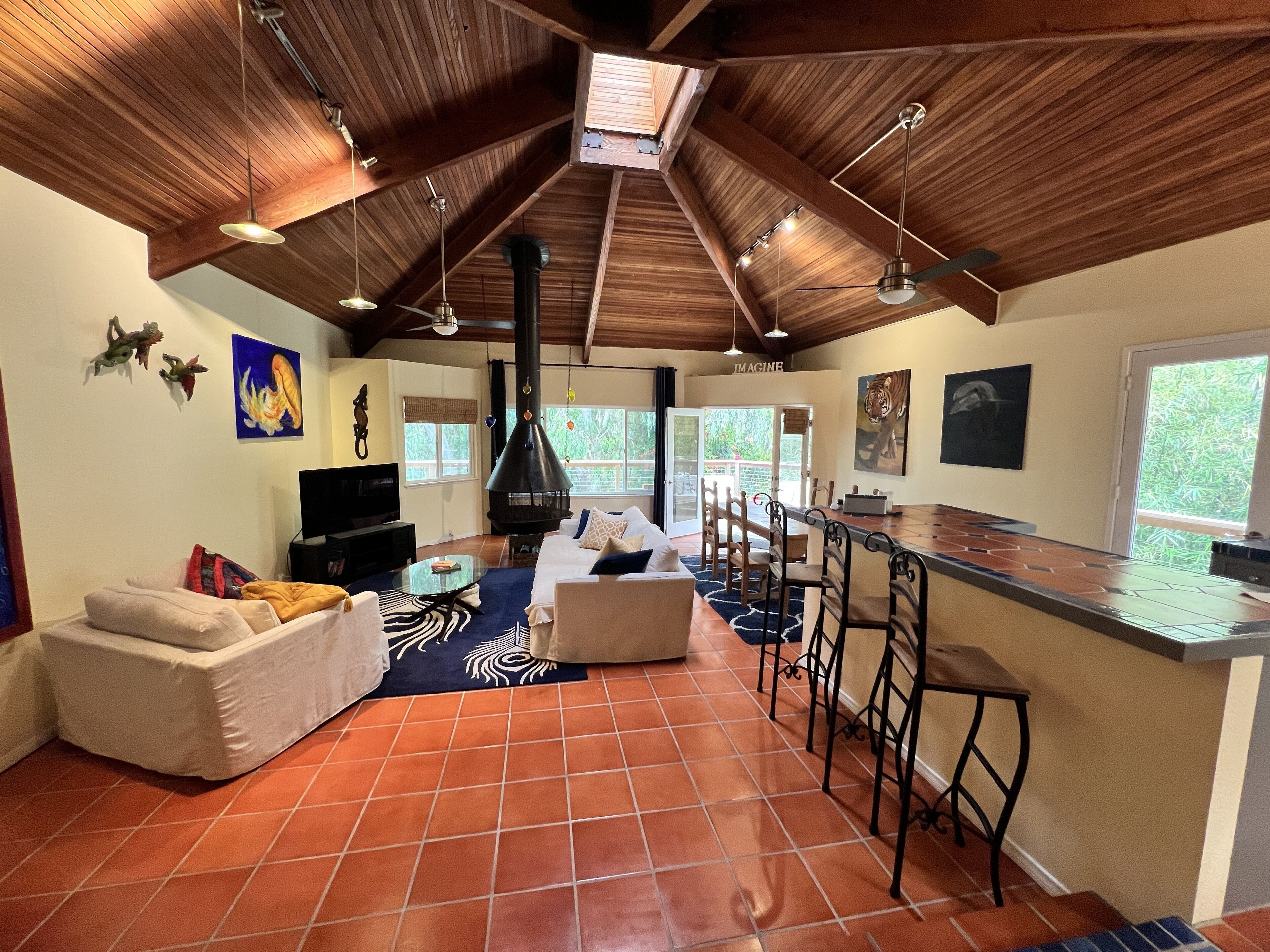 A spacious living area with a high ceiling, a fireplace, a dining area with bar stools, and a large window offering a view of greenery.
