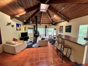 A spacious living area with a high ceiling, a fireplace, a dining area with bar stools, and a large window offering a view of greenery.
