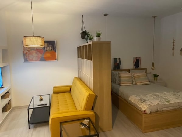 A compact bedroom with a yellow sofa, a wooden bed, a small bedside table, and a hanging plant.