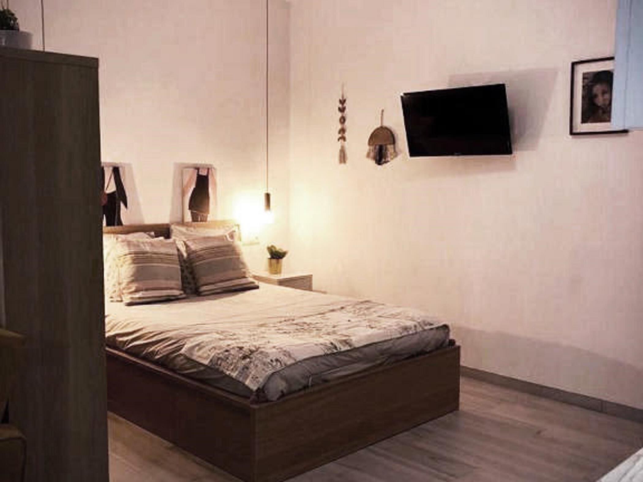 Un dormitorio con cama de madera, una mesita de noche con lámpara, un televisor montado en la pared y un cuadro enmarcado.