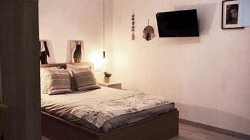 Un dormitorio con cama de madera, una mesita de noche con lámpara, un televisor montado en la pared y un cuadro enmarcado.