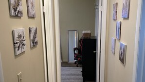 Un couloir aux murs de couleur beige, doté d’un sol recouvert de moquette et orné de plusieurs œuvres d’art encadrées. Une porte donne accès à une autre pièce où se trouvent une chaise et une petite table.