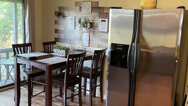 Une cuisine équipée d’un réfrigérateur en acier inoxydable, d’une table à manger en bois, de chaises et d’une fenêtre avec des rideaux.
