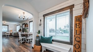 A cozy living room with a wooden bench, a dining area with white chairs, and a large window with blinds. - The Fir Country Cottage (Philomath)